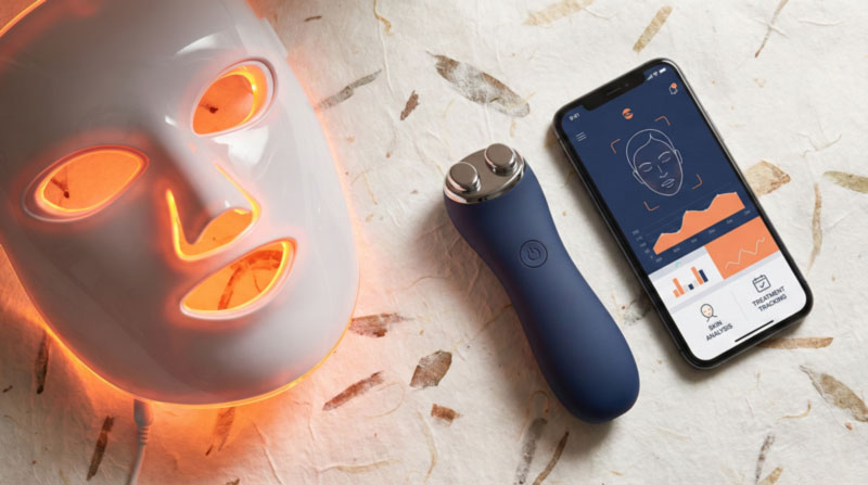 A white LED light therapy face mask glowing with orange light, a dark blue handheld microcurrent facial device with two metal probes, and a smartphone displaying a skincare analysis app with graphs and face scan, all resting on a textured, light-colored paper background with embedded botanical elements.